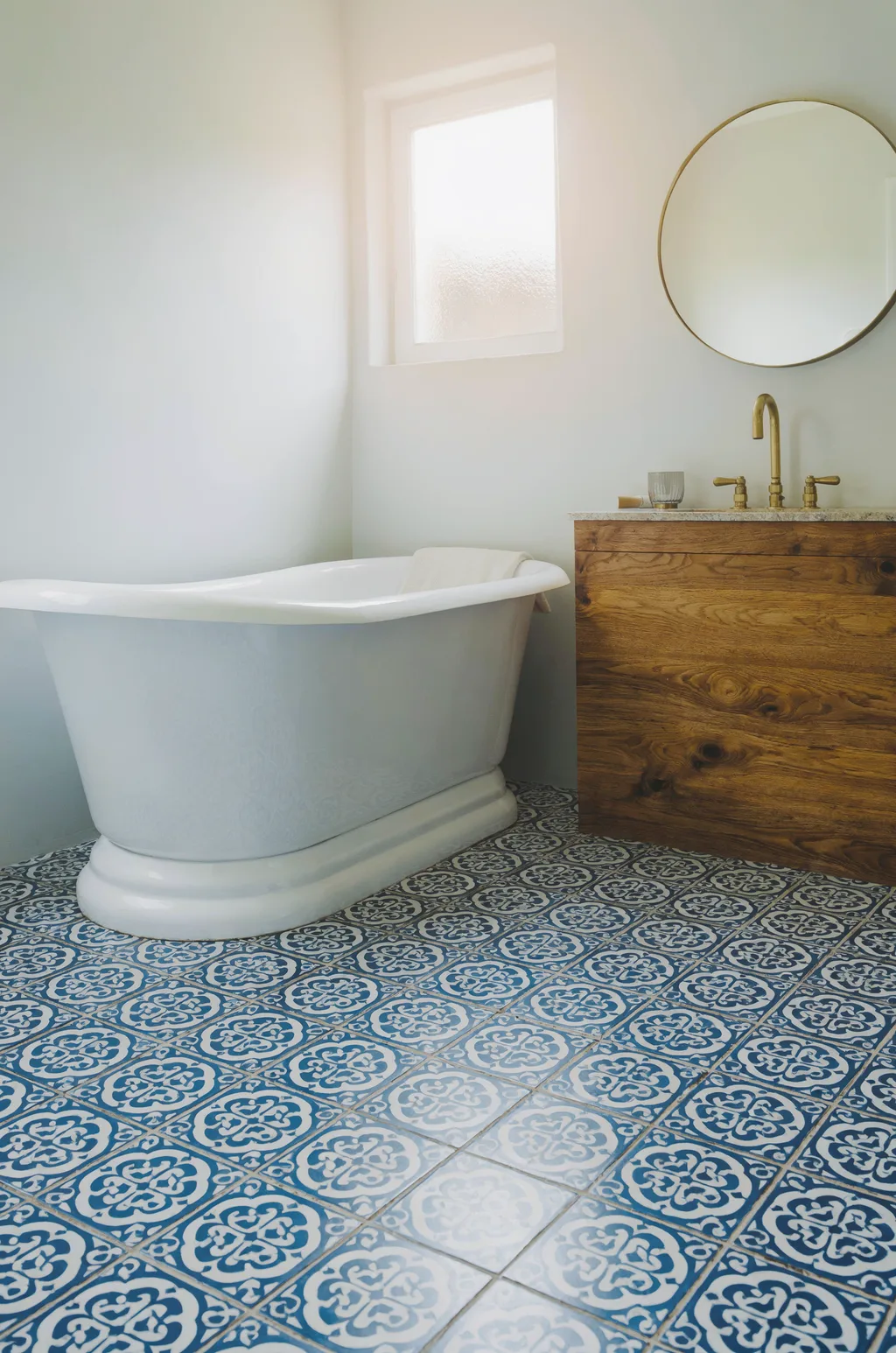 Luma Uni-1 Max: Bathroom interior with blue and white patterned cement floor tiles, a freestanding white bathtub, a round mirror above a wooden vanity and a brass faucet. Morning light from a small frosted window.