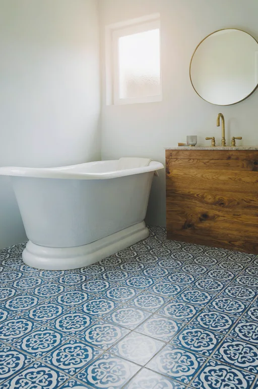 Luma Uni-1 Max: Bathroom interior with blue and white patterned cement floor tiles, a freestanding white bathtub, a round mirror above a wooden vanity and a brass faucet. Morning light from a small frosted window.
