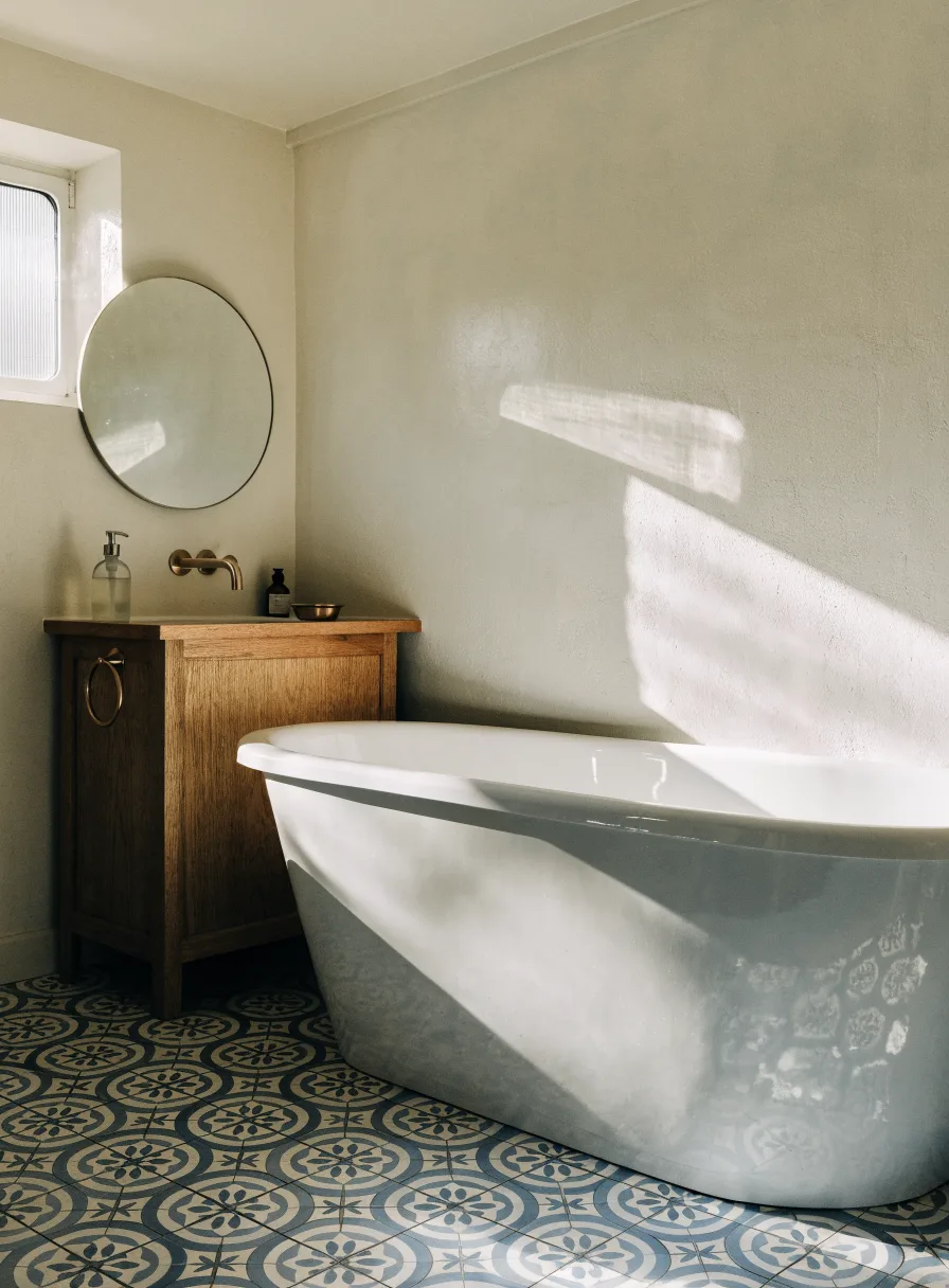 Recraft V4.1: Bathroom interior with blue and white patterned cement floor tiles, a freestanding white bathtub, a round mirror above a wooden vanity and a brass faucet. Morning light from a small frosted window.