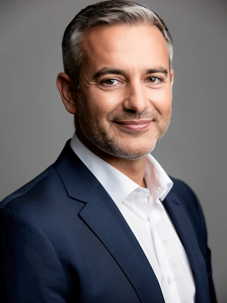 FLUX.2 [pro]: Professional corporate headshot of a man in his forties with short salt-and-pepper hair, wearing a navy suit and a white shirt, with a slight smile, against a neutral mid-gray seamless backdrop. Soft key light from the left with a gentle fill, sharp focus on the eyes, shallow depth of field.