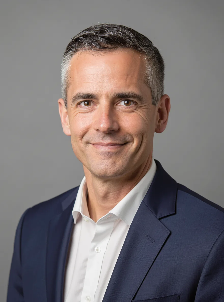FLUX 3: Professional corporate headshot of a man in his forties with short salt-and-pepper hair, wearing a navy suit and a white shirt, with a slight smile, against a neutral mid-gray seamless backdrop. Soft key light from the left with a gentle fill, sharp focus on the eyes, shallow depth of field.