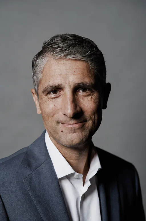 Luma Uni-1 Max: Professional corporate headshot of a man in his forties with short salt-and-pepper hair, wearing a navy suit and a white shirt, with a slight smile, against a neutral mid-gray seamless backdrop. Soft key light from the left with a gentle fill, sharp focus on the eyes, shallow depth of field.