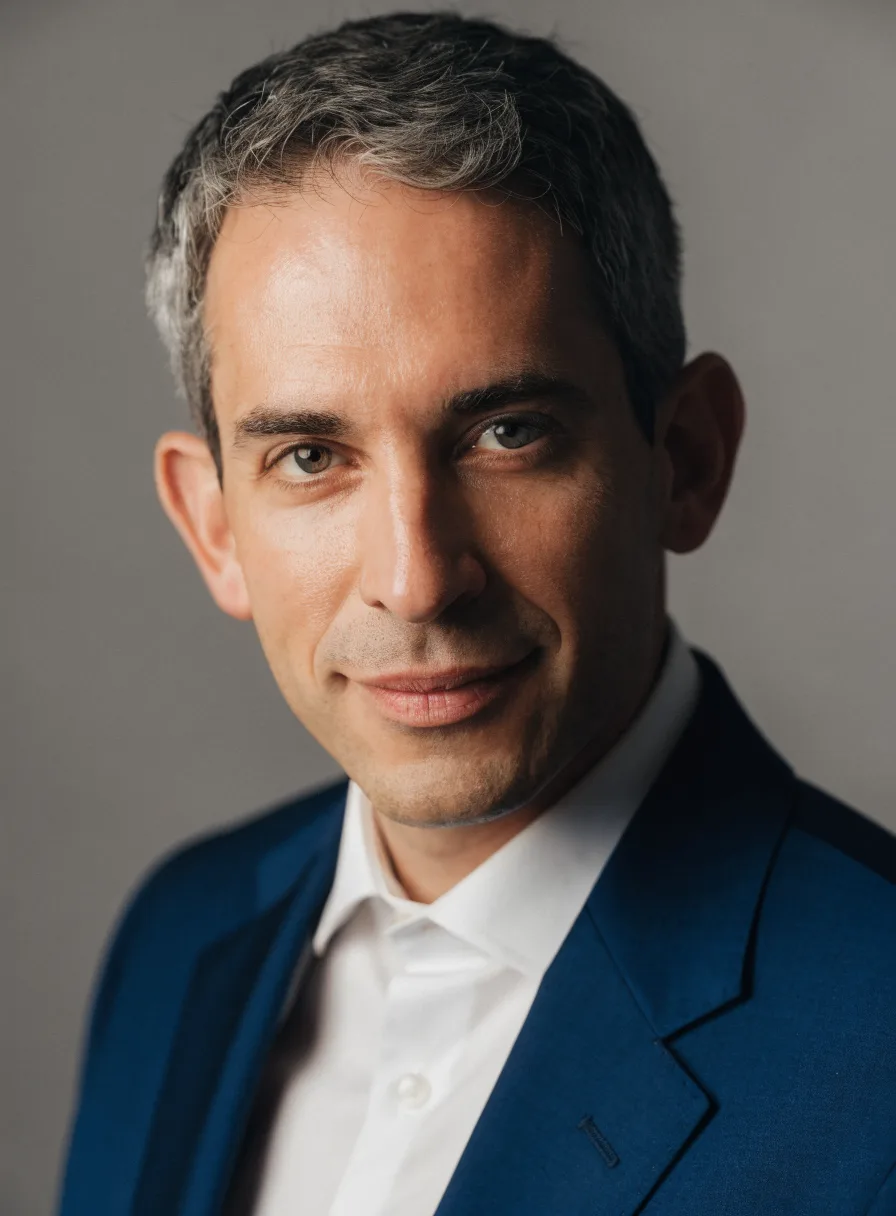 Recraft V4.1: Professional corporate headshot of a man in his forties with short salt-and-pepper hair, wearing a navy suit and a white shirt, with a slight smile, against a neutral mid-gray seamless backdrop. Soft key light from the left with a gentle fill, sharp focus on the eyes, shallow depth of field.