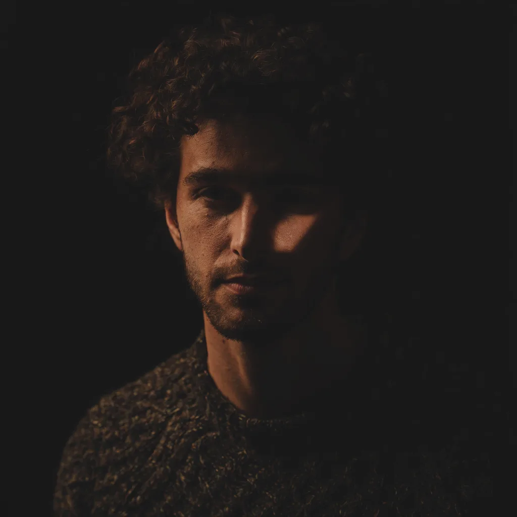 Luma Uni-1 Max: Fine-art portrait of a young man with curly dark hair and a short beard, wearing a textured wool sweater. Rembrandt lighting with a small triangle of light on the shadow-side cheek, a black background, moody low-key photographic look.