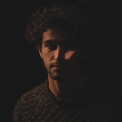 Luma Uni-1 Max: Fine-art portrait of a young man with curly dark hair and a short beard, wearing a textured wool sweater. Rembrandt lighting with a small triangle of light on the shadow-side cheek, a black background, moody low-key photographic look.