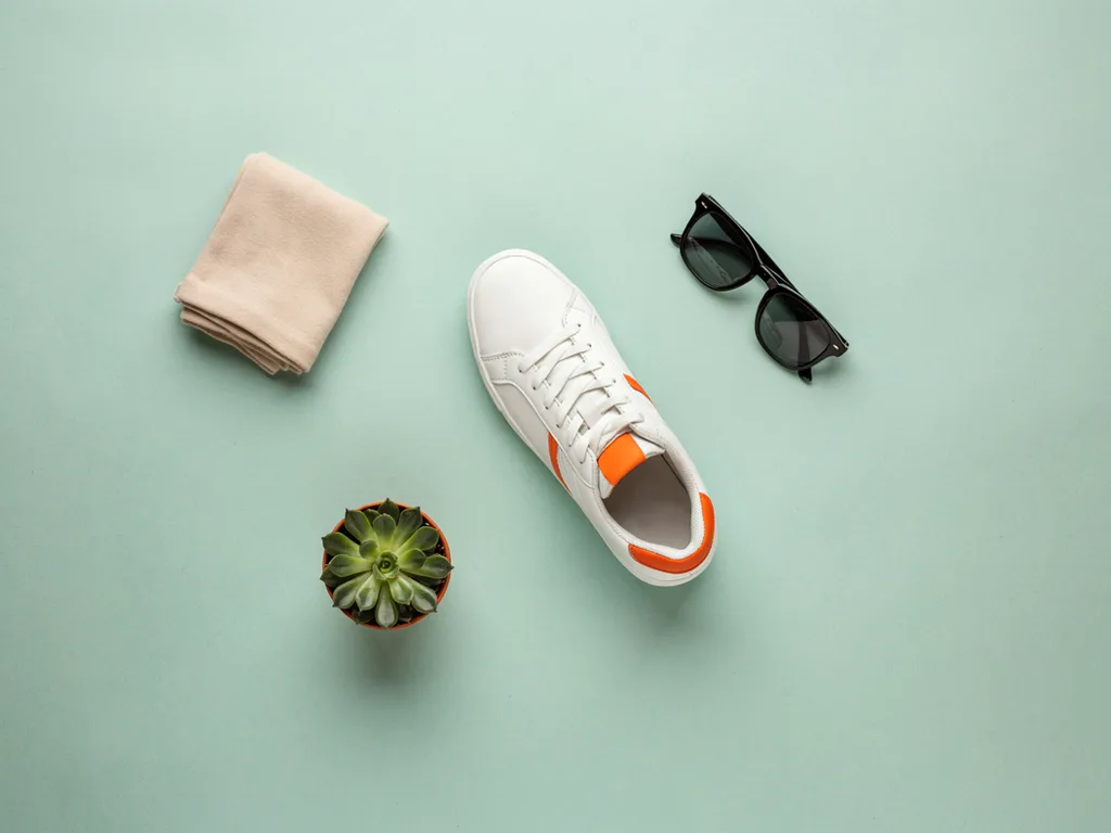 Ideogram V4.5: Top-down flat lay e-commerce photo of a single white leather sneaker with orange accents on a pastel sage green background, surrounded by a folded beige sock, a pair of sunglasses and a small potted succulent. Even soft lighting, crisp short shadows, generous empty space.