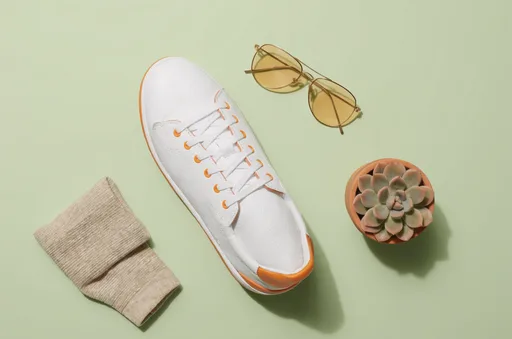 Luma Uni-1 Max: Top-down flat lay e-commerce photo of a single white leather sneaker with orange accents on a pastel sage green background, surrounded by a folded beige sock, a pair of sunglasses and a small potted succulent. Even soft lighting, crisp short shadows, generous empty space.