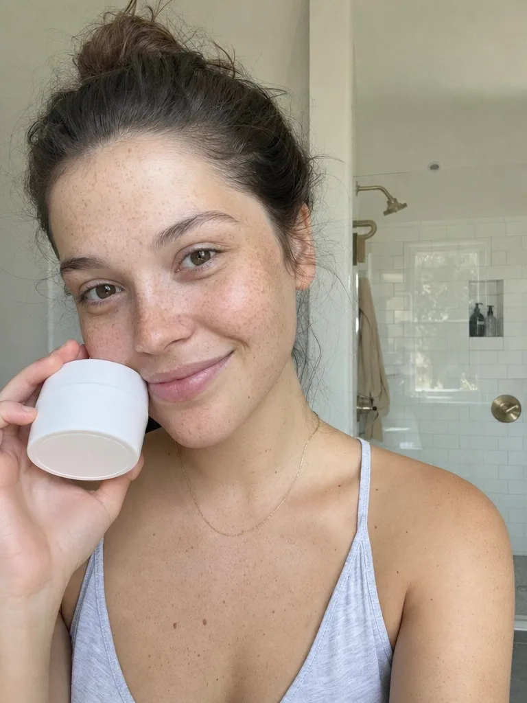 GPT Image 2: Authentic user-generated style photo for a skincare ad: a smiling young woman with freckles holds a small plain white moisturizer jar next to her cheek in a bright bathroom, natural daylight, a slight phone-camera look, no heavy makeup.