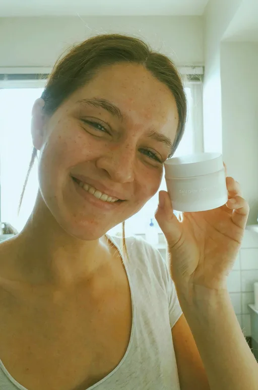 Luma Uni-1 Max: Authentic user-generated style photo for a skincare ad: a smiling young woman with freckles holds a small plain white moisturizer jar next to her cheek in a bright bathroom, natural daylight, a slight phone-camera look, no heavy makeup.